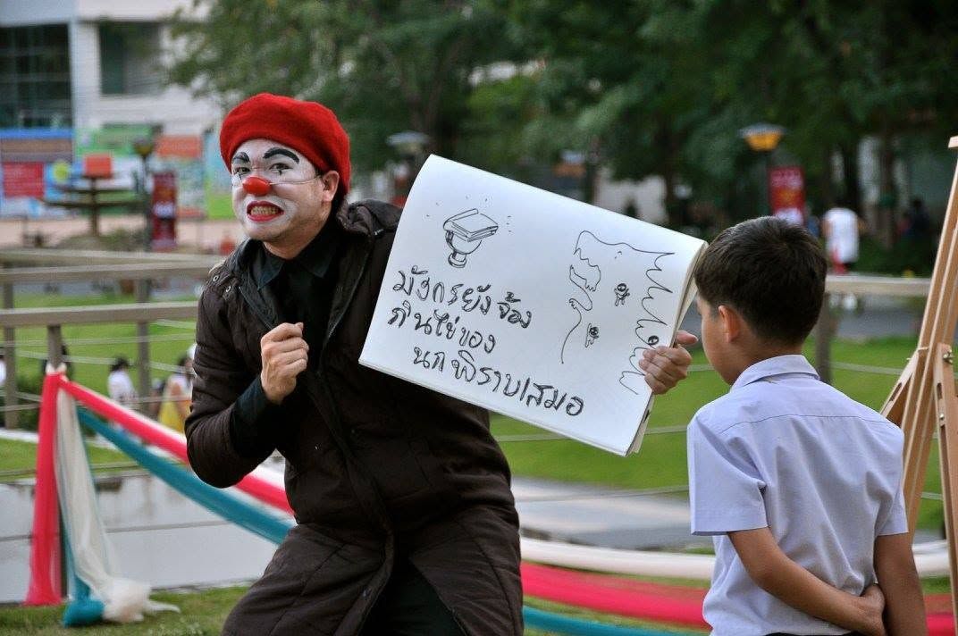 Wasu as a clown in a red hat holding up a hand-drawn sign for a schoolboy outdoors